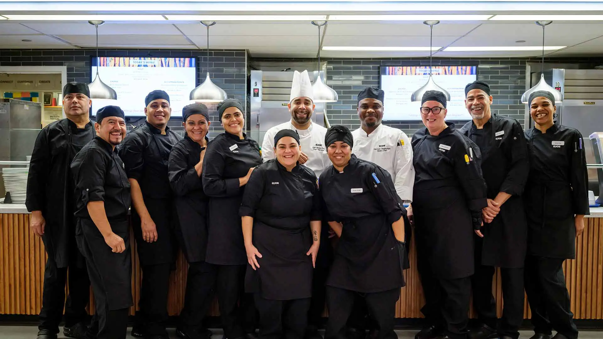 A diverse group of eleven chefs and kitchen staff, dressed in black uniforms and white chef hats, pose together and smile confidently in a modern commercial kitchen.