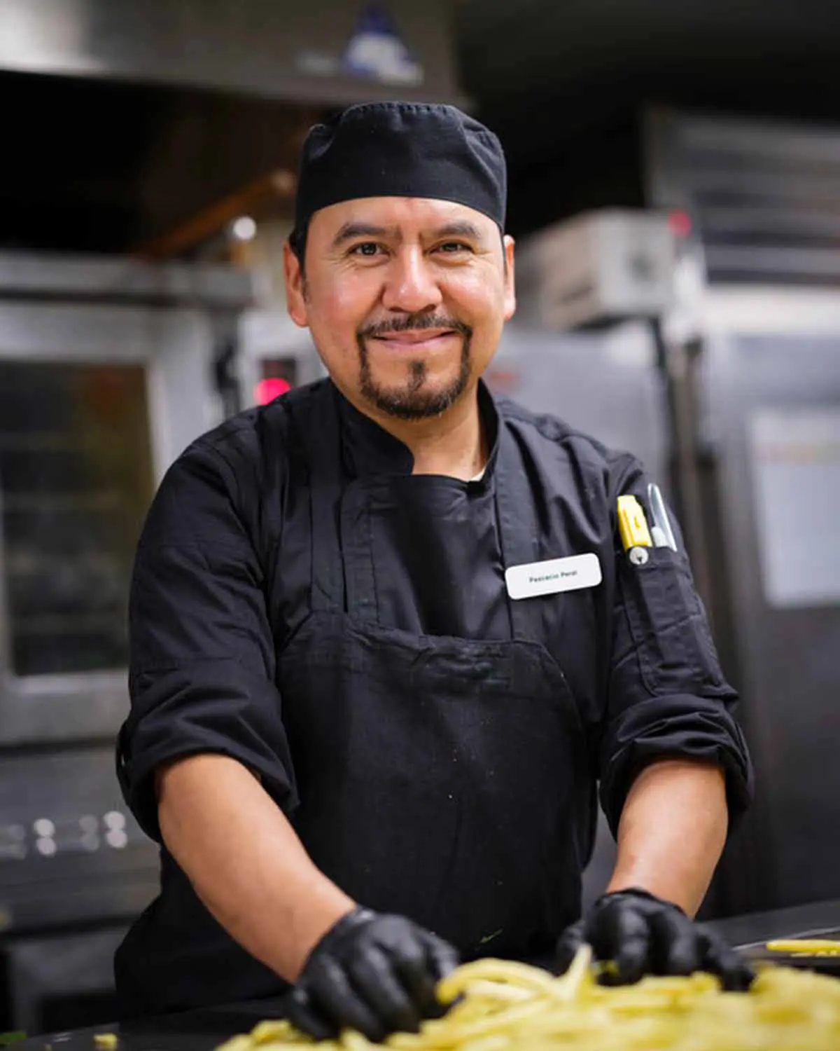 A chef wearing black attire and gloves, smiling while preparing food in a commercial kitchen.