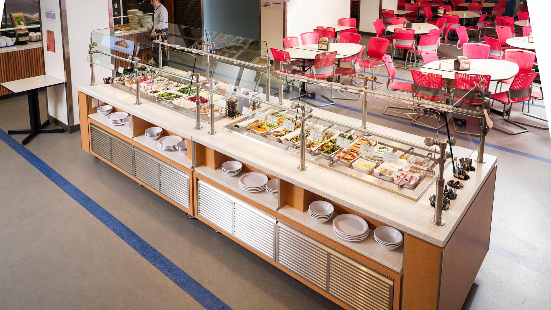 A modern cafeteria with a self-service food counter displaying various dishes, while a person stands behind the counter and rows of red chairs and tables are arranged in the background.